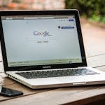 A MacBook Pro displaying Google Search on a wooden table outdoors, next to a smartphone.