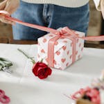 A person wrapping a heart-patterned gift with a red ribbon, accompanied by a rose.