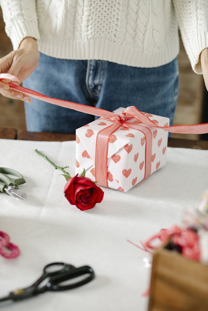 A person wrapping a heart-patterned gift with a red ribbon, accompanied by a rose.